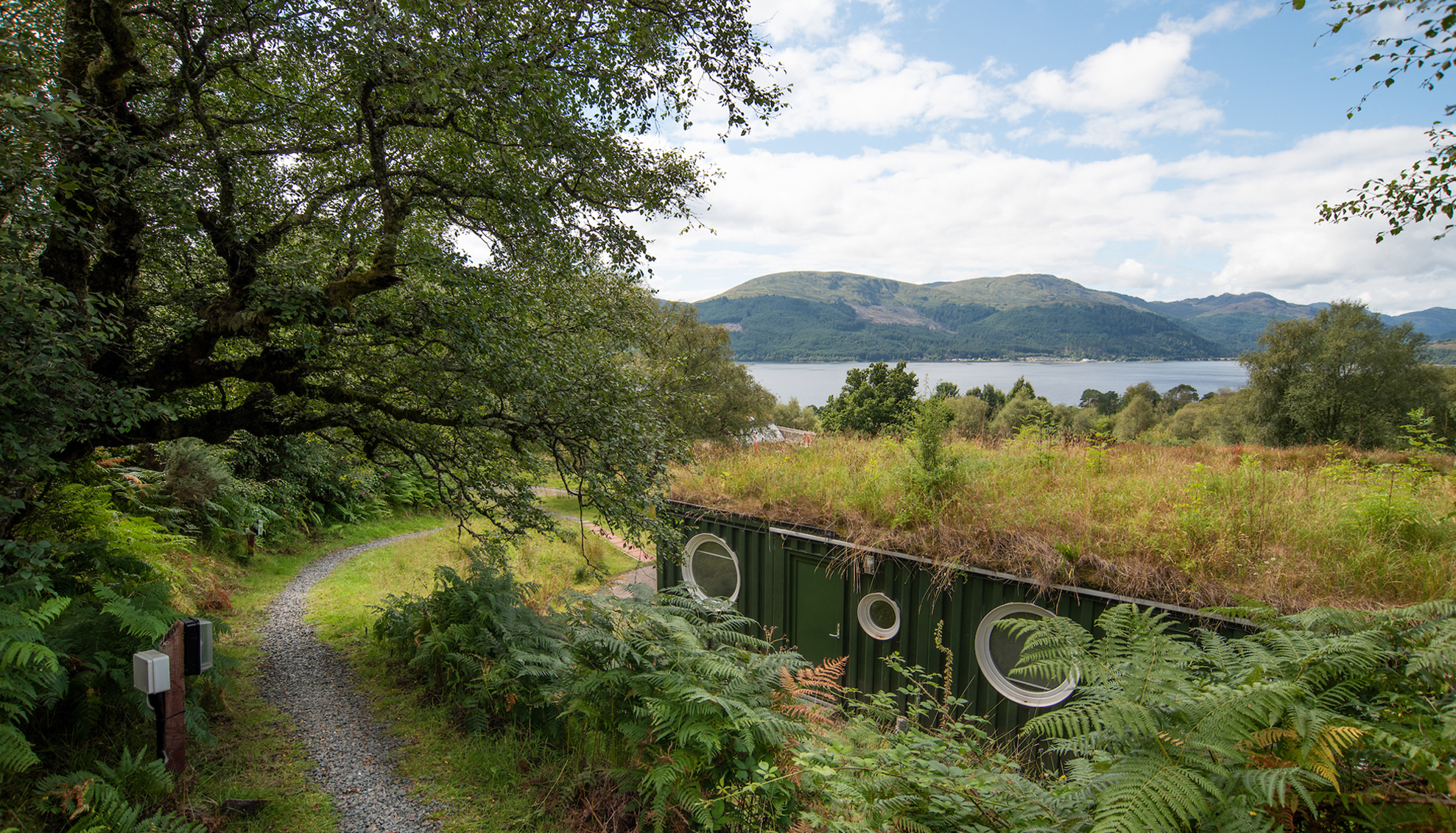 The view of Cove Park's Cubes - dark green metal shipping containers converted into units of accommodation, with porthole windows framed in white - from the path which cuts across Cove Park's site. The image is framed to the left with a mature tree in full leaf. The blue waters of Lock Long and the grey and green hills of Cowal Peninsula are visible in the distance.