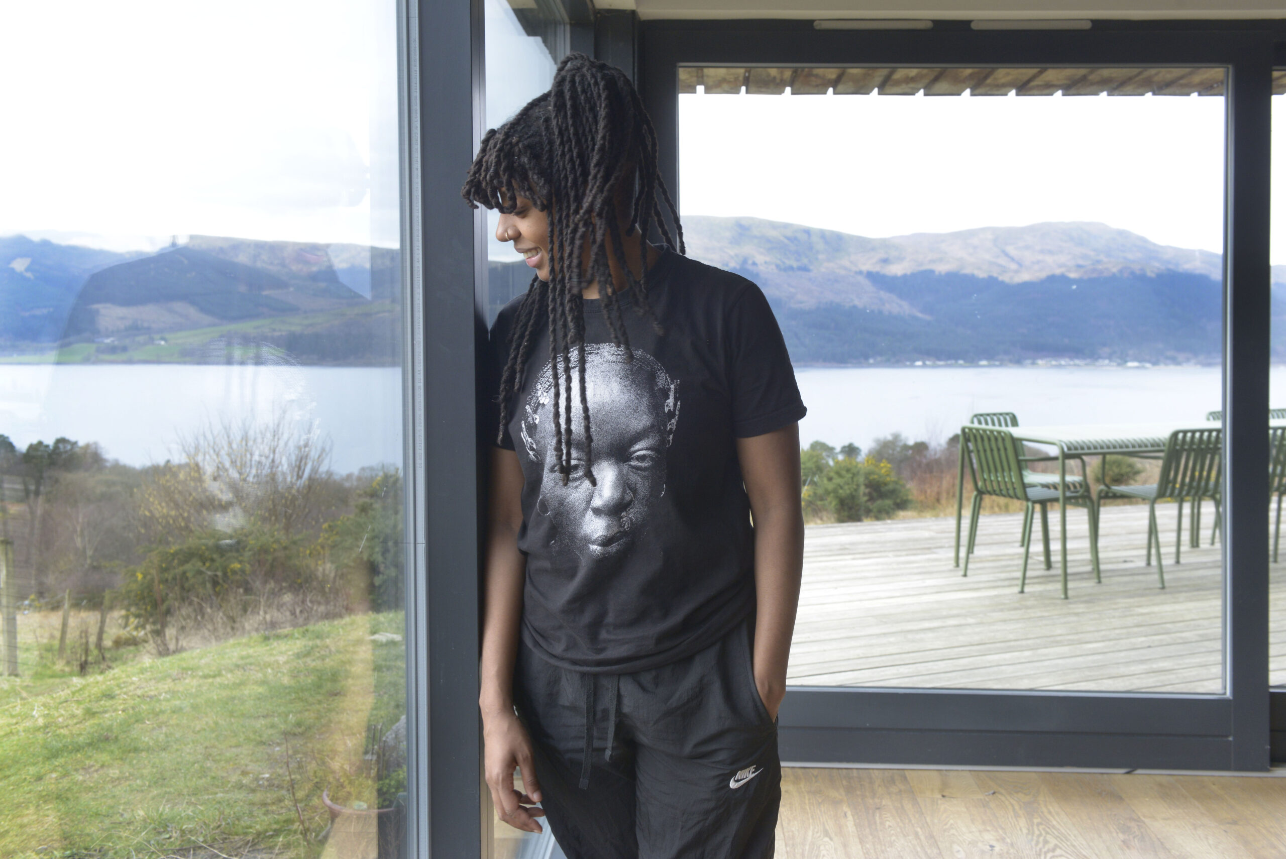 A Black woman wearing a protective, braided hairstyle, leans against a large glass window and looks out toward her shoulder. Through the windows, you can see a lush, green landscape, Loch Long, and the hills of Cowal Peninsula.