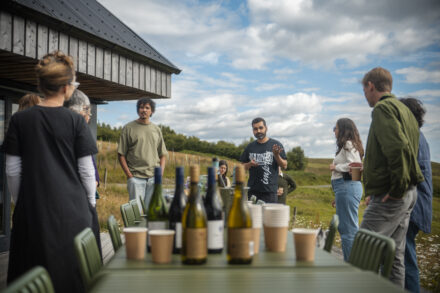 two people talking standing at the end of a long outdoor table. There are wine bottles and various cups on the table in front of them.