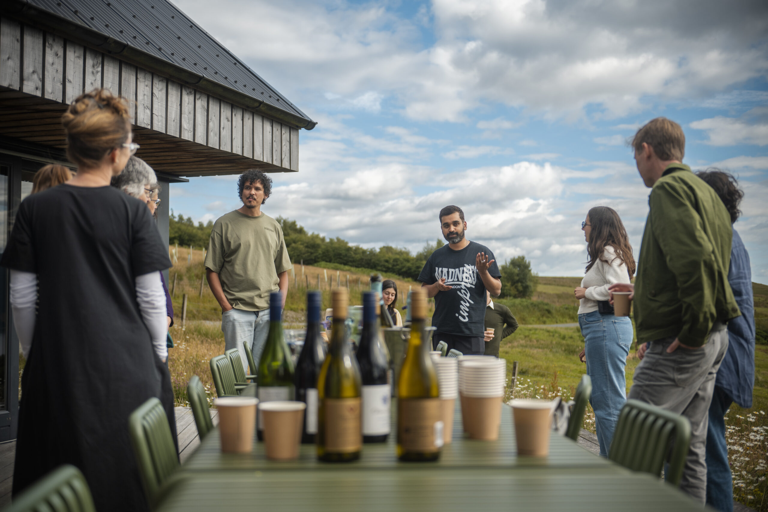 two people talking standing at the end of a long outdoor table. There are wine bottles and various cups on the table in front of them.