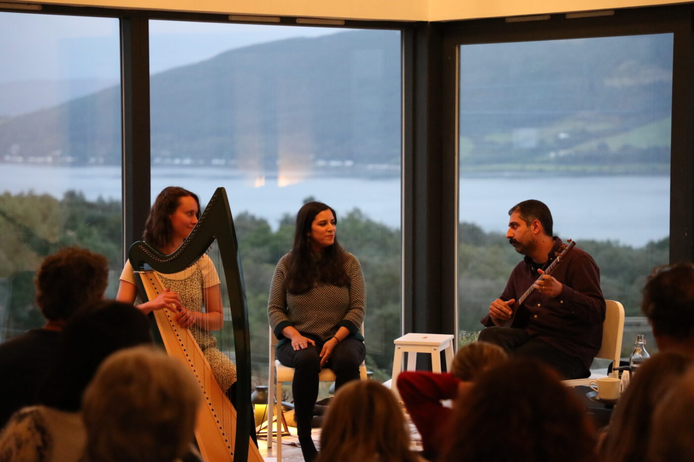 Three musicians perform for a group of people. One of the musicians is playing a traditional guitar and another plays a harp. They are sitting in front of a large corner window overlooking Loch Long and the hills of the Cowal Peninsula at dusk.