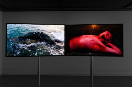 An exhibition space with two flat video screens side by side. On the left is an image of a rocky beach with waves, on the right is an image of a shirtless Trans man in a red glow.