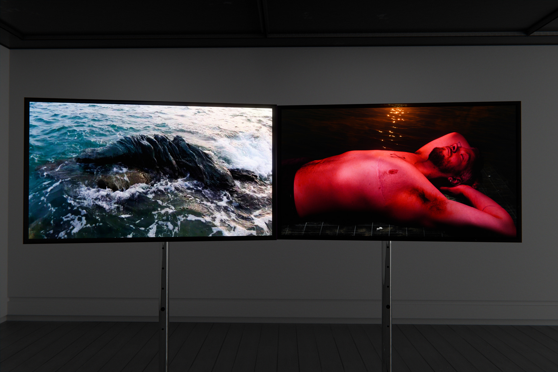 An exhibition space with two flat video screens side by side. On the left is an image of a rocky beach with waves, on the right is an image of a shirtless Trans man in a red glow.