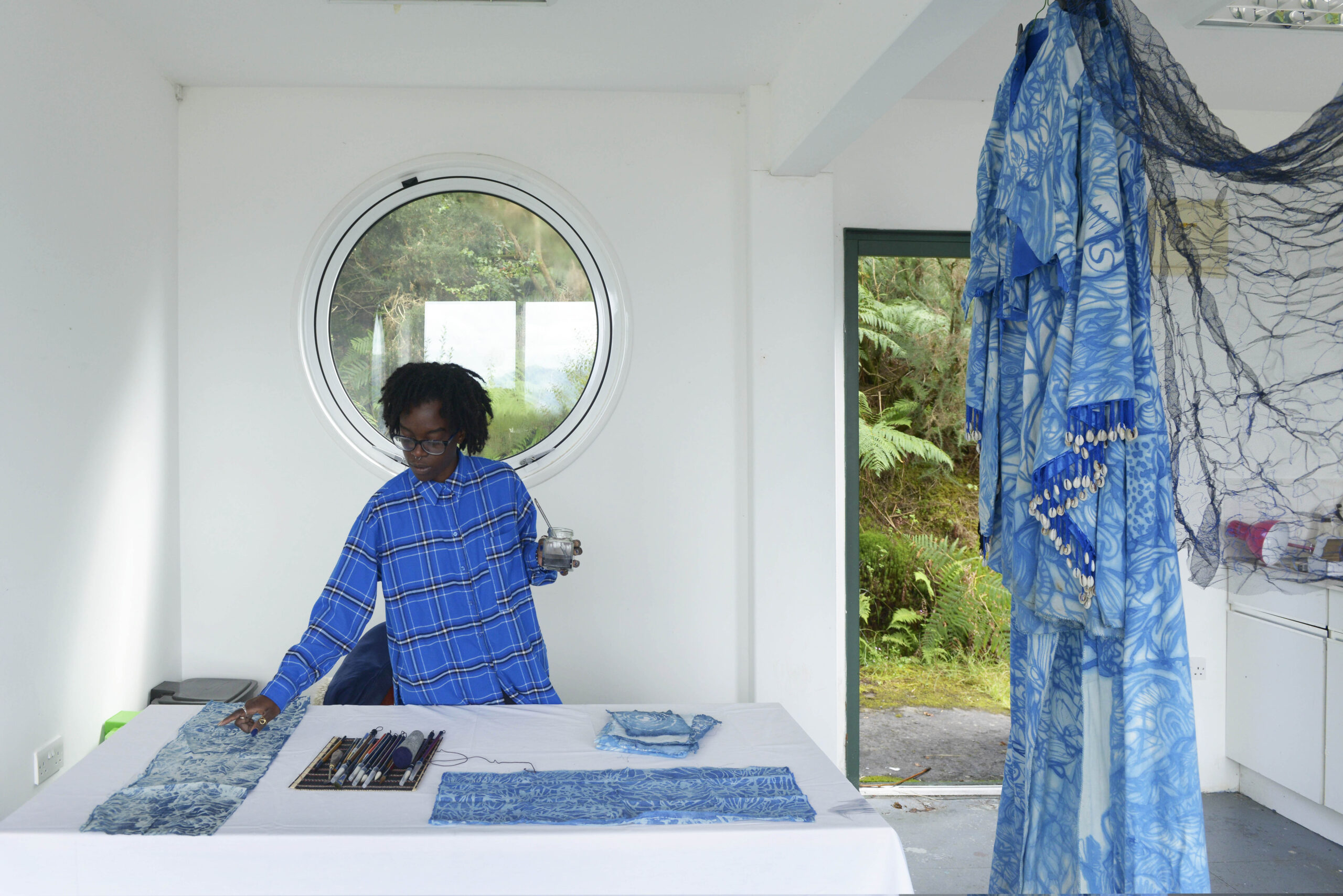 A Black woman wearing a cobalt blue flannel shirt paints in her studio with various other types of blue. There is a chiffon gown, in bright turquoise blue, hanging on the side.