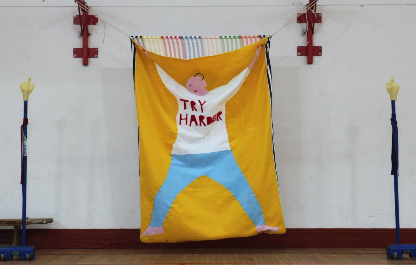 Image of artwork – a large textile hung from a white wall. It is mustard yellow and features the outline of a person. The words 'Try Harder' are written on the figure's shirt.